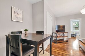A dining area with a wooden table and chairs, a television on a wooden stand, and a door leading to a bedroom with a bed and window. - Luxury Condo down the street from Tower Grove Park (St. Louis)