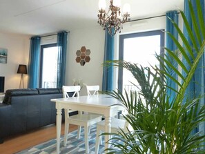 Un salon comprenant un canapé, une table à manger avec des chaises, un lustre et une grande fenêtre ornée de rideaux bleus.
