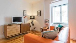 Una sala moderna con un aparador de madera, un televisor, una lámpara de pie, un sofá y una ventana con vistas a edificios.