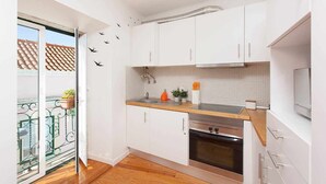 Cocina compacta con armarios blancos, encimera de madera, horno integrado y ventana con vistas a un tejado con baldosas.