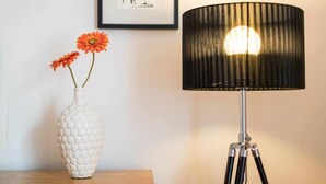 Un jarrón hexagonal blanco con flores anaranjadas sobre una superficie de madera, una lámpara con borde negro y bombilla blanca sobre un atril, y un cuadro en la pared.