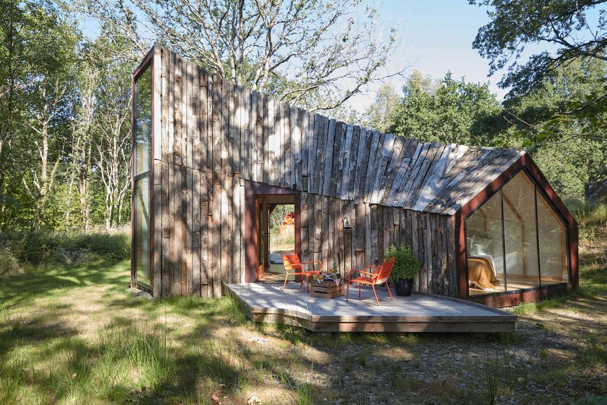 A rustic cabin with a large glass window, a wooden deck, and a cozy seating area.