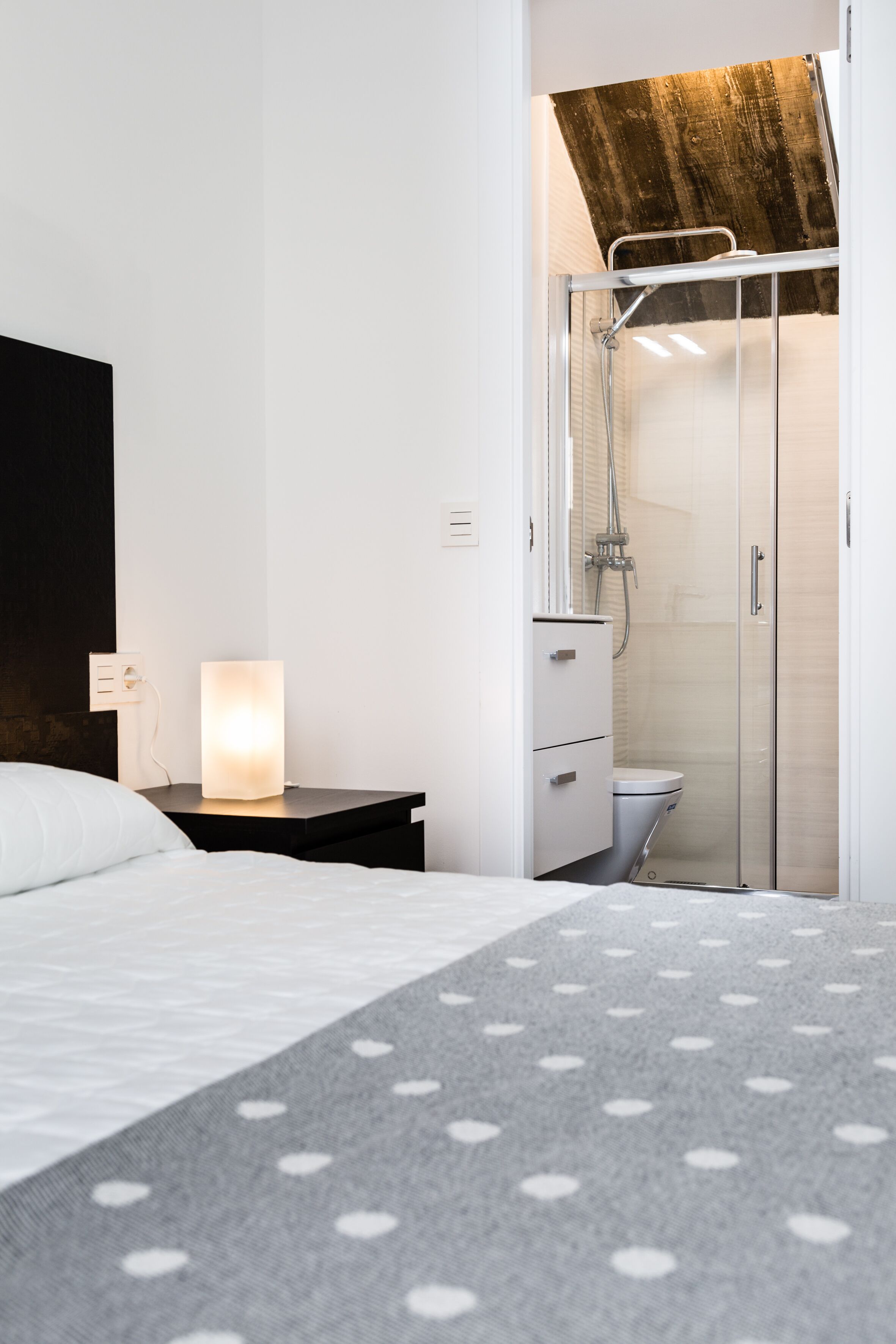A modern hotel room with a bed, a bedside table with a lamp, a shower area, and a wardrobe.