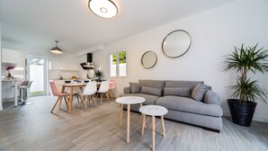 Ein modernes Wohnzimmer mit einem grauen Sofa, zwei runden weißen Beistelltischen, einem Essbereich mit einem weißen Tisch und rosafarbenen Stühlen sowie einer großen Topfpflanze.