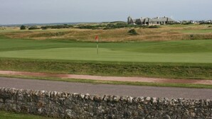 En golfbane med en stenvæg i forgrunden og en bygning med en unik arkitektonisk stil i baggrunden.