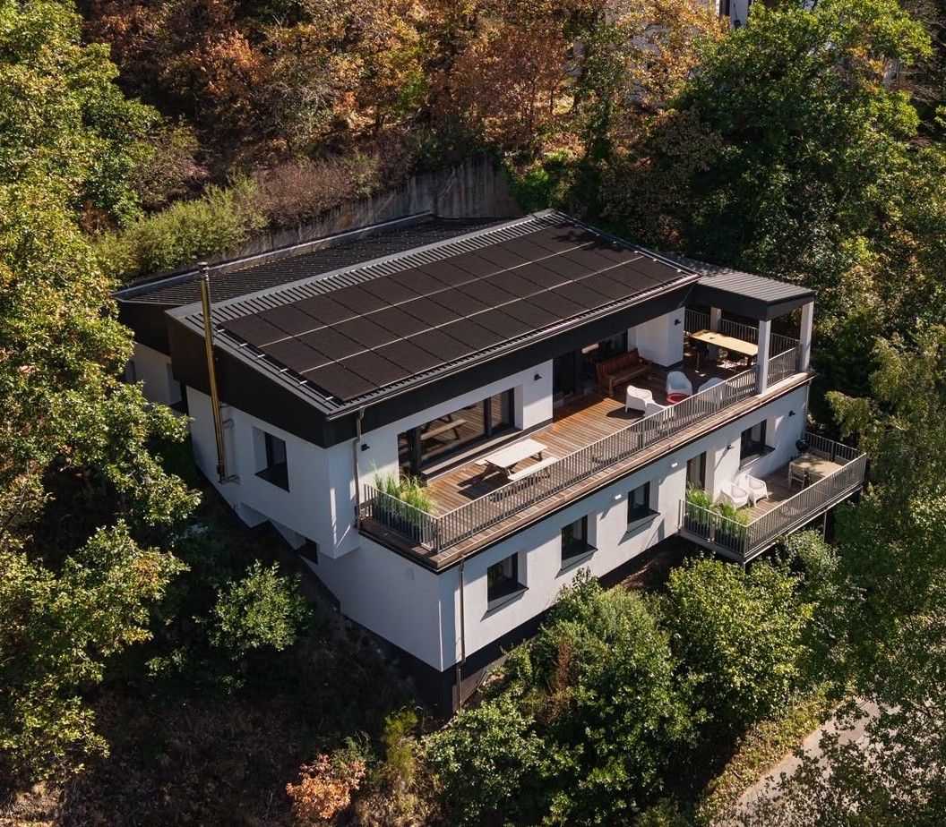 A modern two-story house with a flat roof, large windows, and a balcony with outdoor furniture.