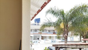 A covered outdoor area with a car parked under a wooden structure, a palm tree, and a building with multiple floors in the background.