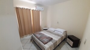 A bedroom with a bed, curtains, an air conditioning unit, a bedside table, and a dresser.