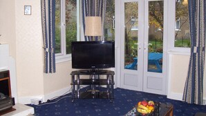 Un salon avec un téléviseur posé sur un meuble, une lampe et une table sur laquelle se trouvent des fruits.