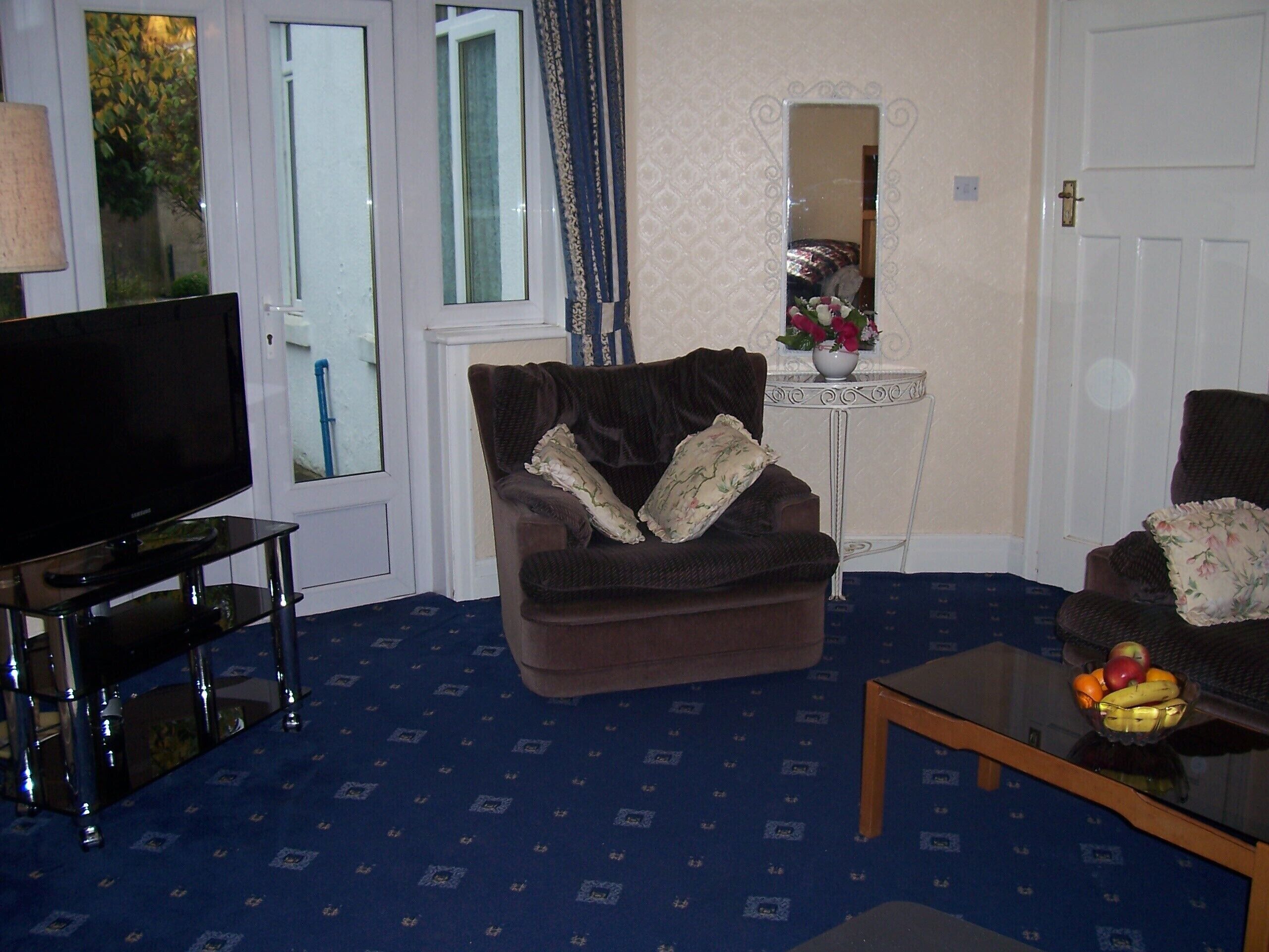 A living room with a dark brown sofa, a television on a glass stand, a small table with fruit, and a mirror on the wall.