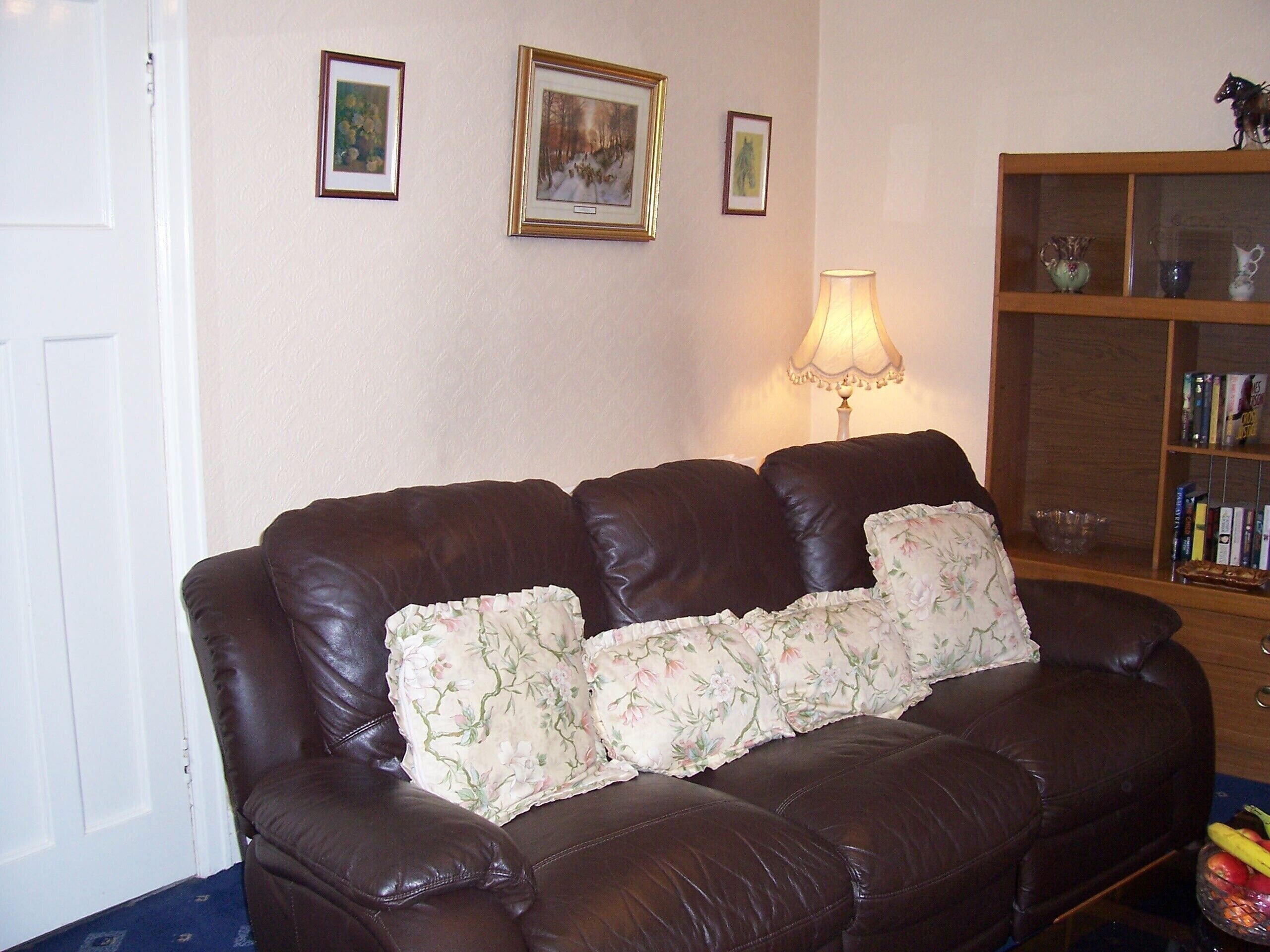 A brown leather sofa with floral cushions, a lamp, and a bookshelf with books and decorative items.