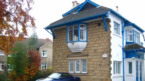 Une maison à deux étages avec une façade en pierre et des éléments décoratifs bleus, une voiture garée devant, et le numéro « 153 » sur le mur.