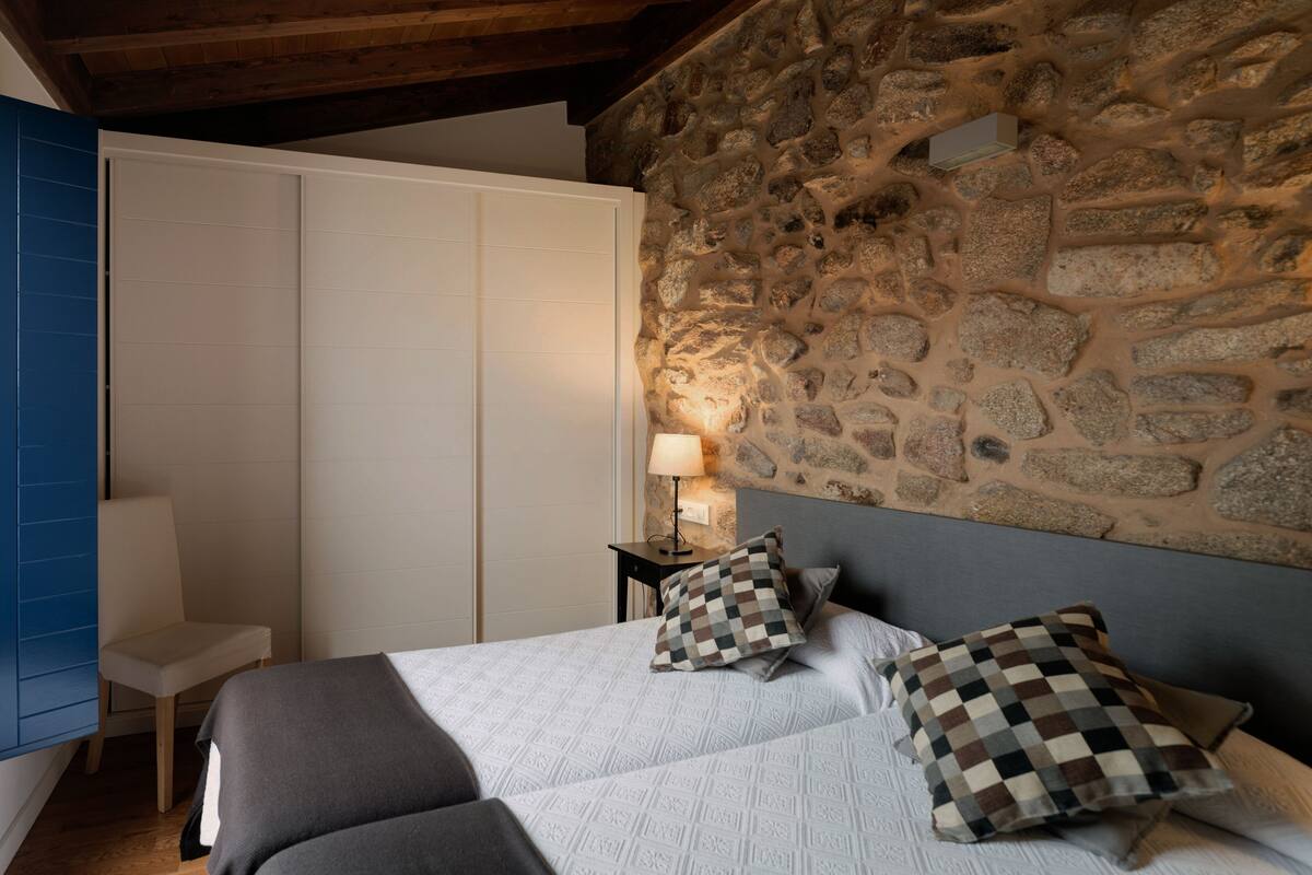 A bedroom with a stone wall, a bed with patterned bedding, a chair, and a nightstand with a lamp.