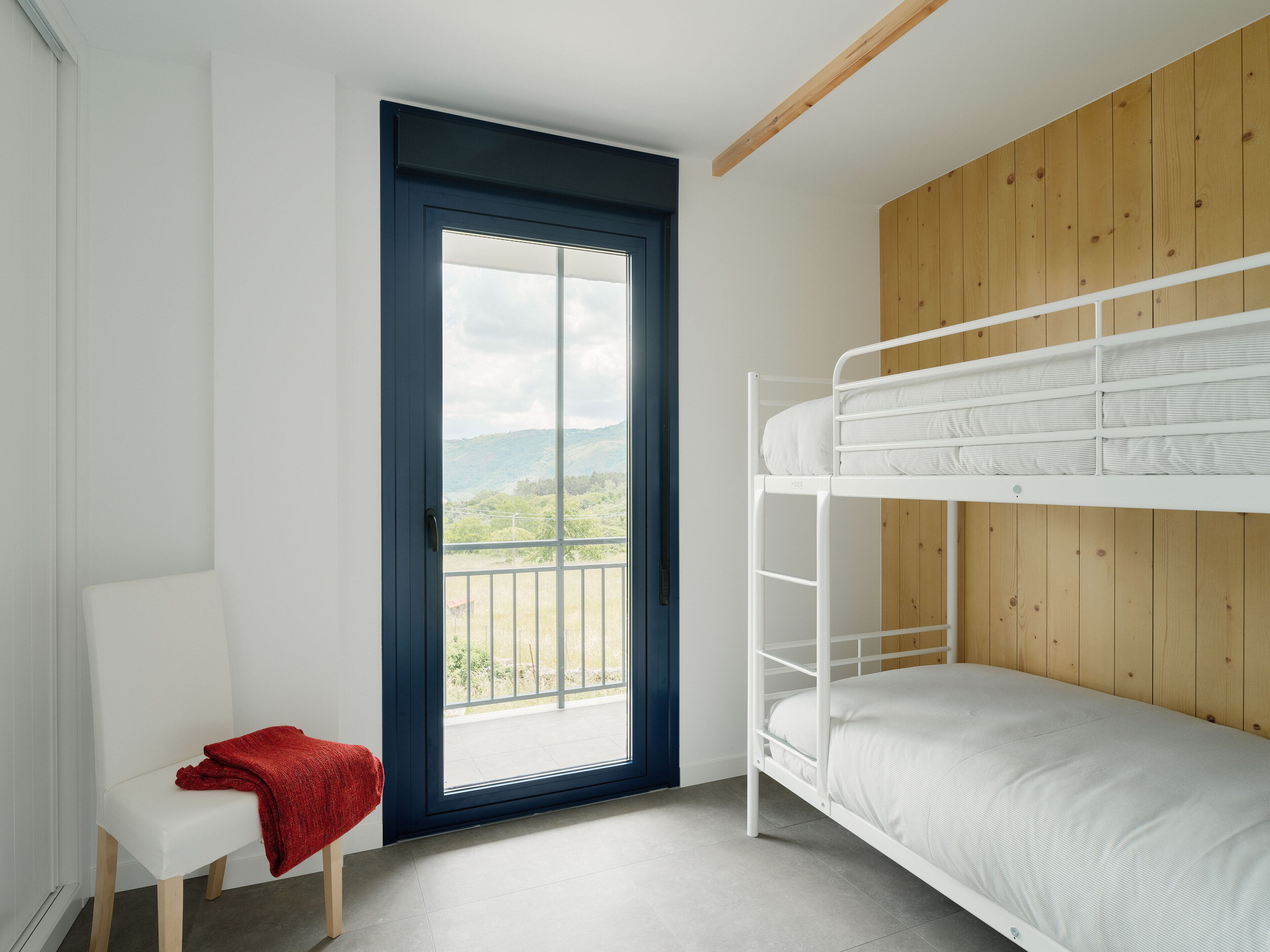 Une pièce avec un lit superposé, une petite chaise blanche recouverte d’un plaid rouge et une porte donnant sur un balcon avec vue sur les collines.