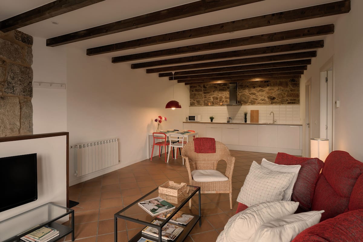 A spacious living area with a modern kitchen in the background, featuring exposed wooden beams and a stone accent wall.