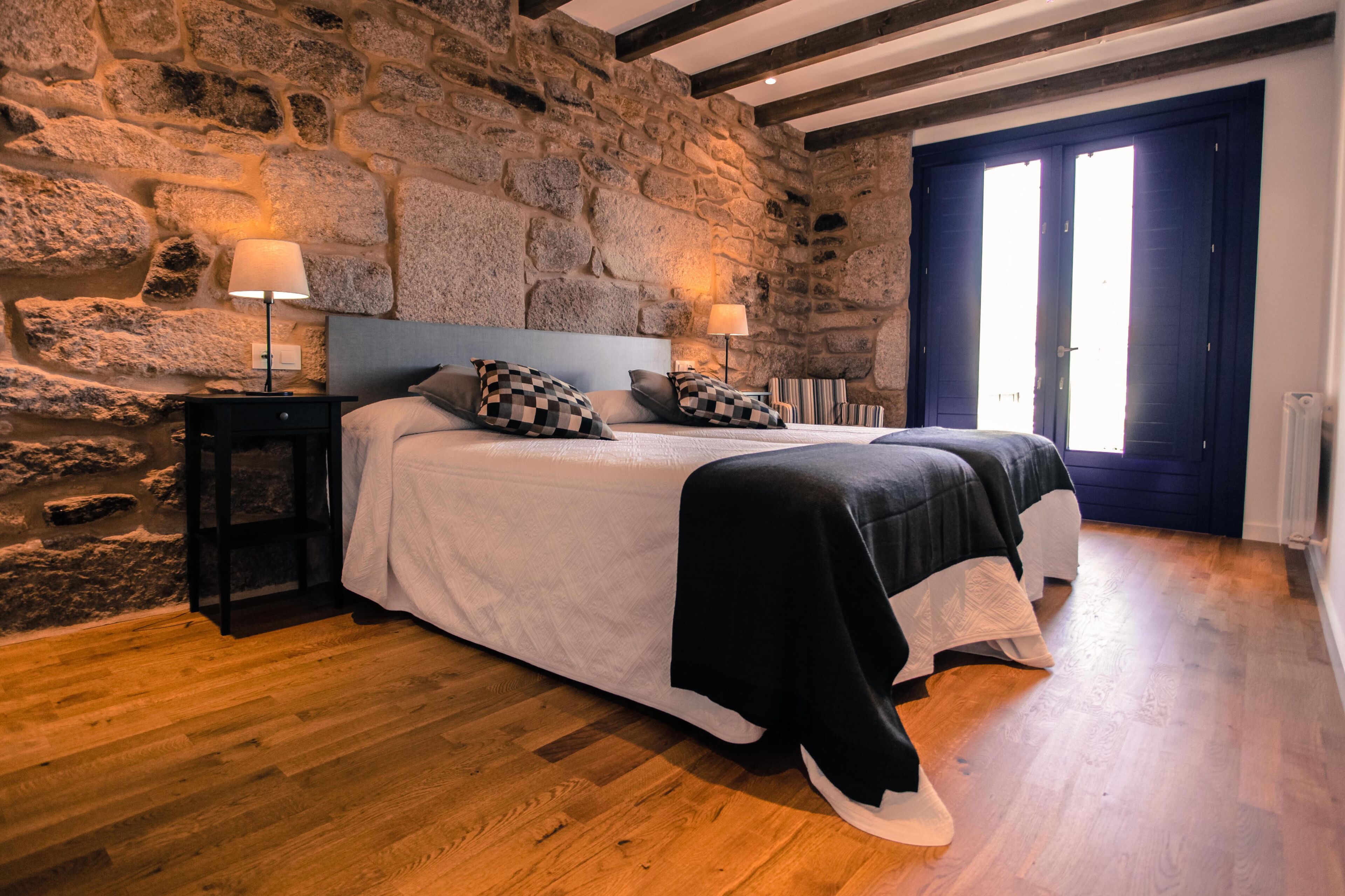 A bedroom with a stone wall, a double bed with black and white bedding, two bedside tables with lamps, and a large window with a view.