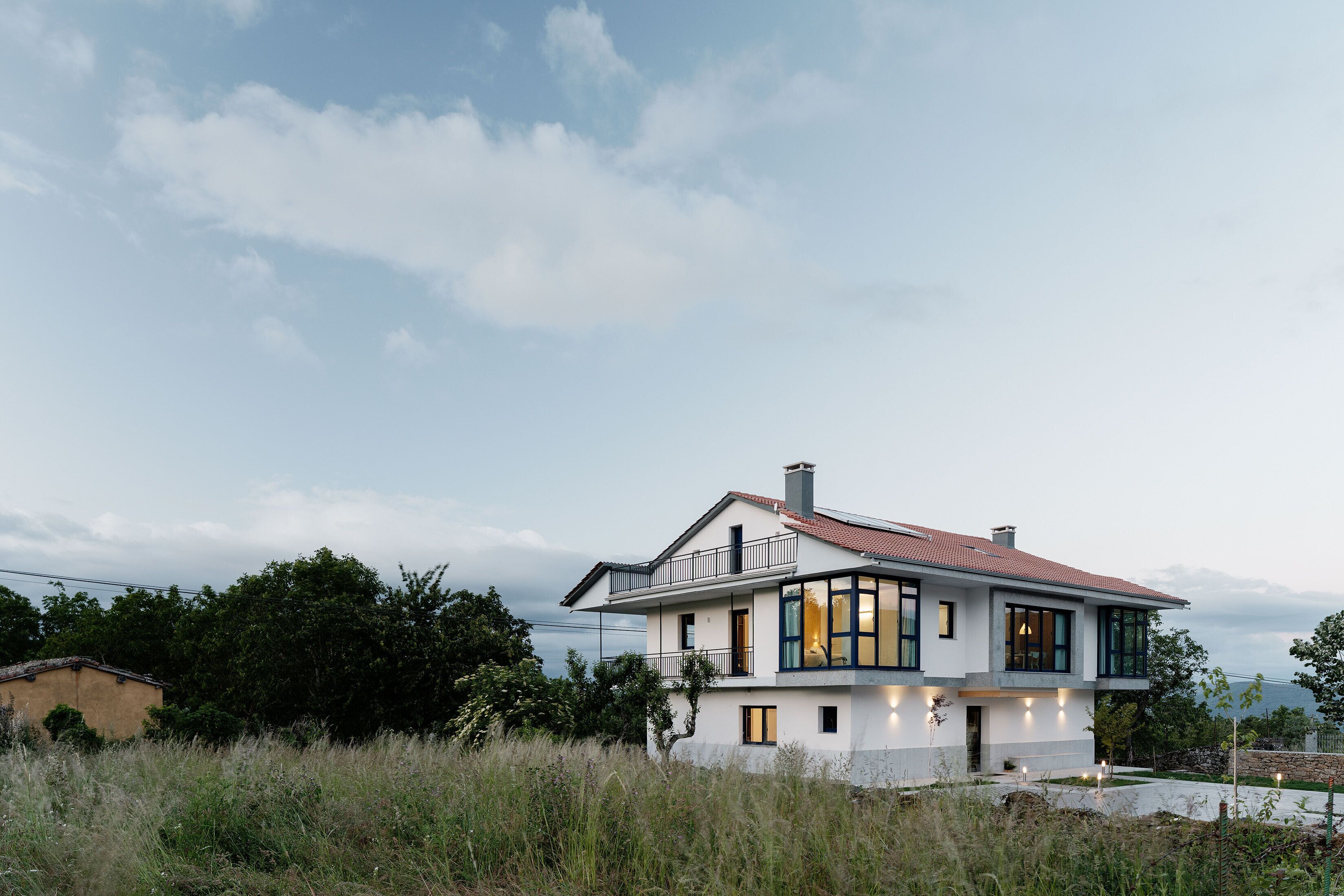 A modern two-story house with a balcony, large windows, and a stone wall in the background.