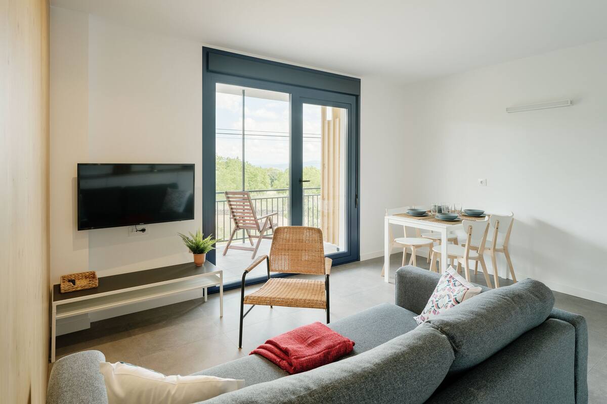 A modern living room with a flat-screen TV, a grey sofa, a dining table with chairs, and a balcony with a view of greenery.