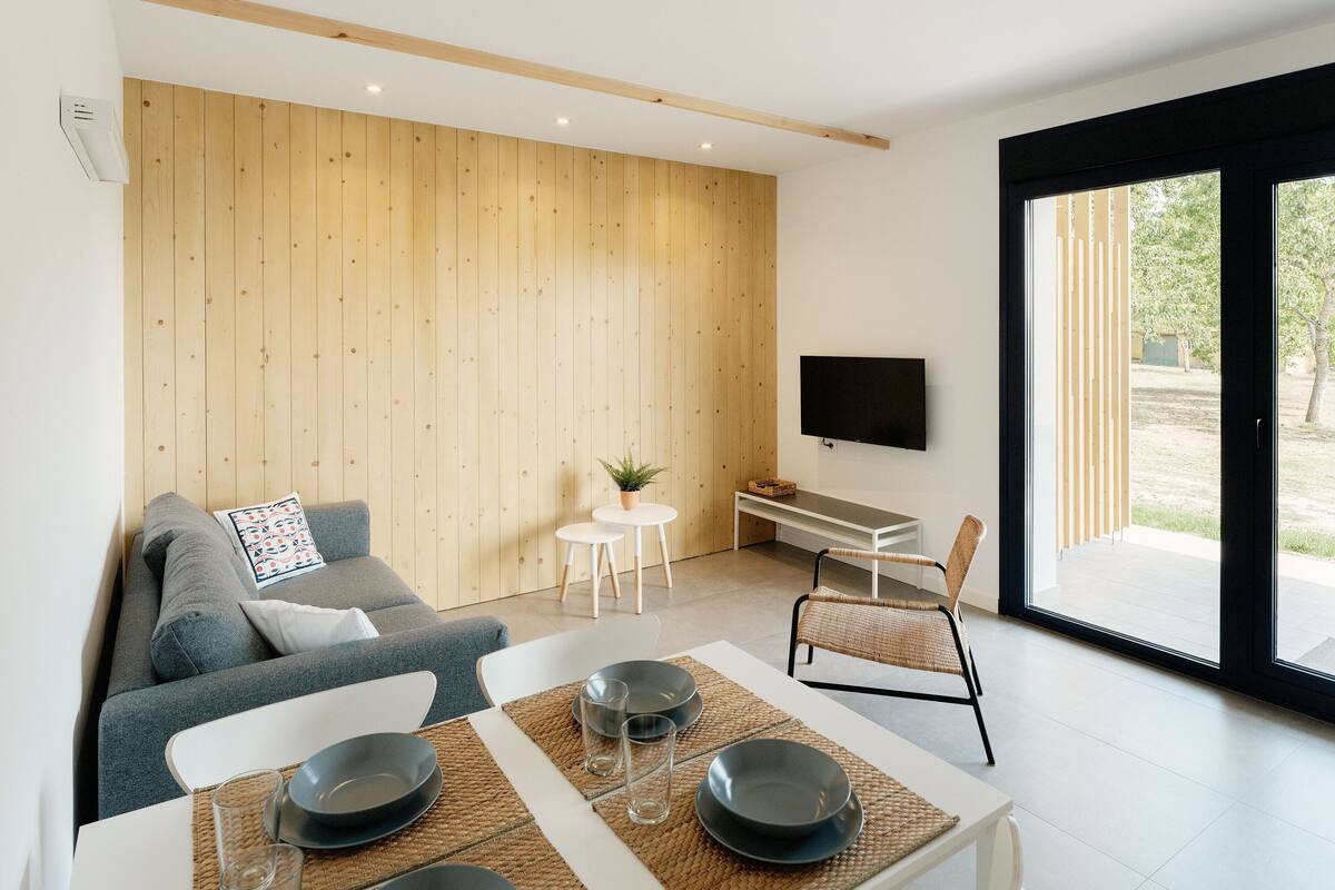 A modern living room with a sofa, a dining table set with plates and glasses, a small round table with a plant, and a TV mounted on the wall.