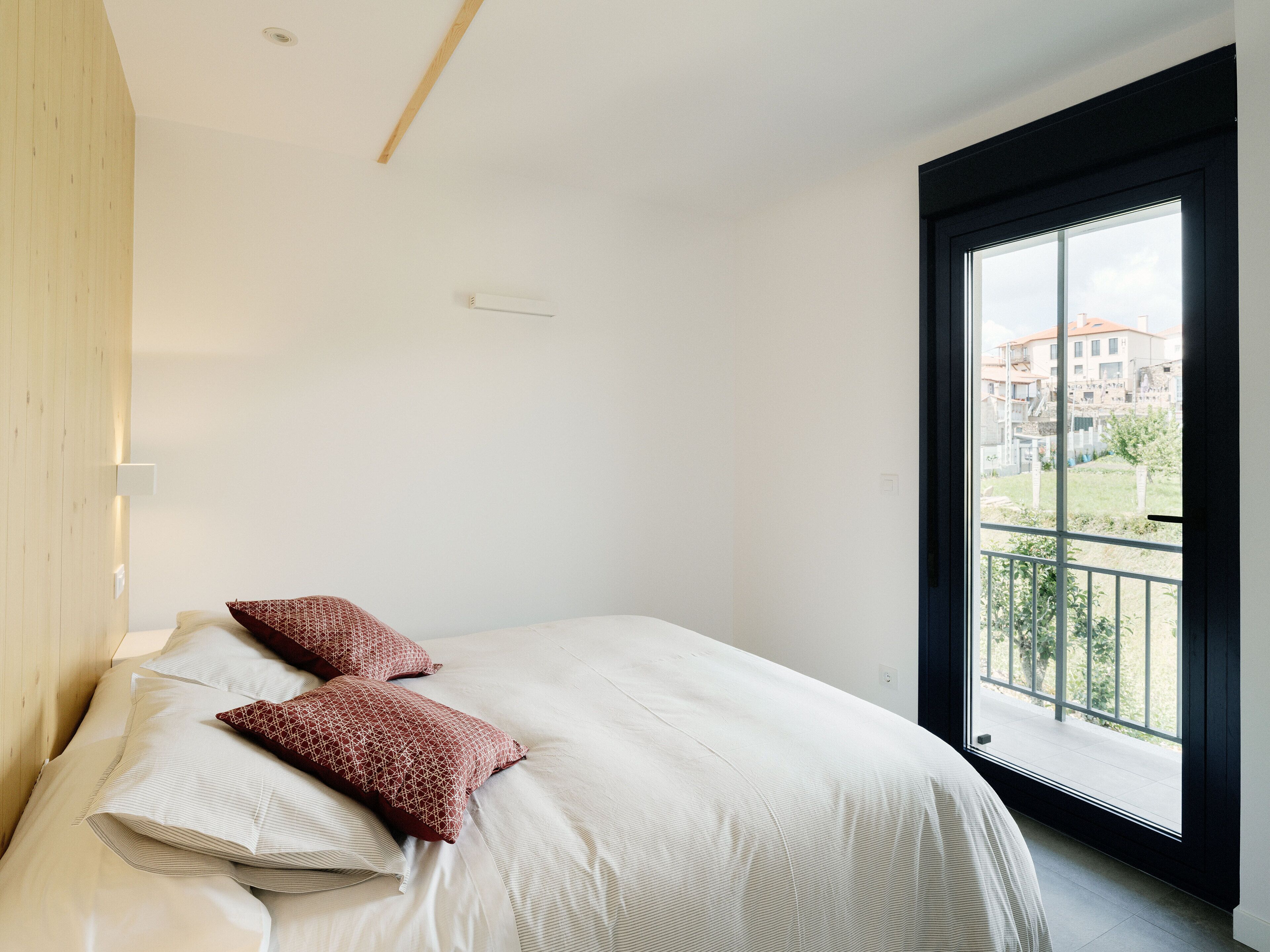 A bedroom with a large bed, two pillows, a wooden headboard, a window with a view of buildings, and a balcony.
