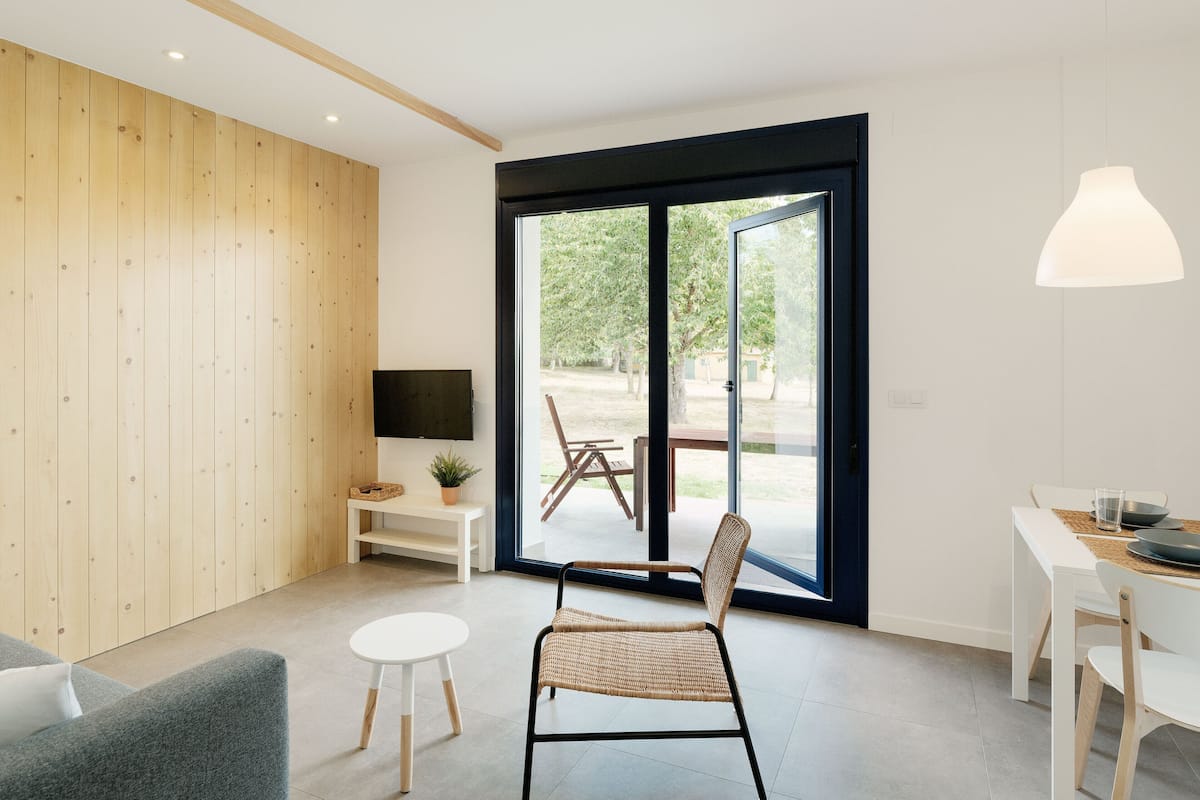 A modern living room with a large sliding glass door leading to an outdoor area, a flat-screen TV mounted on the wall, and a dining table with chairs.