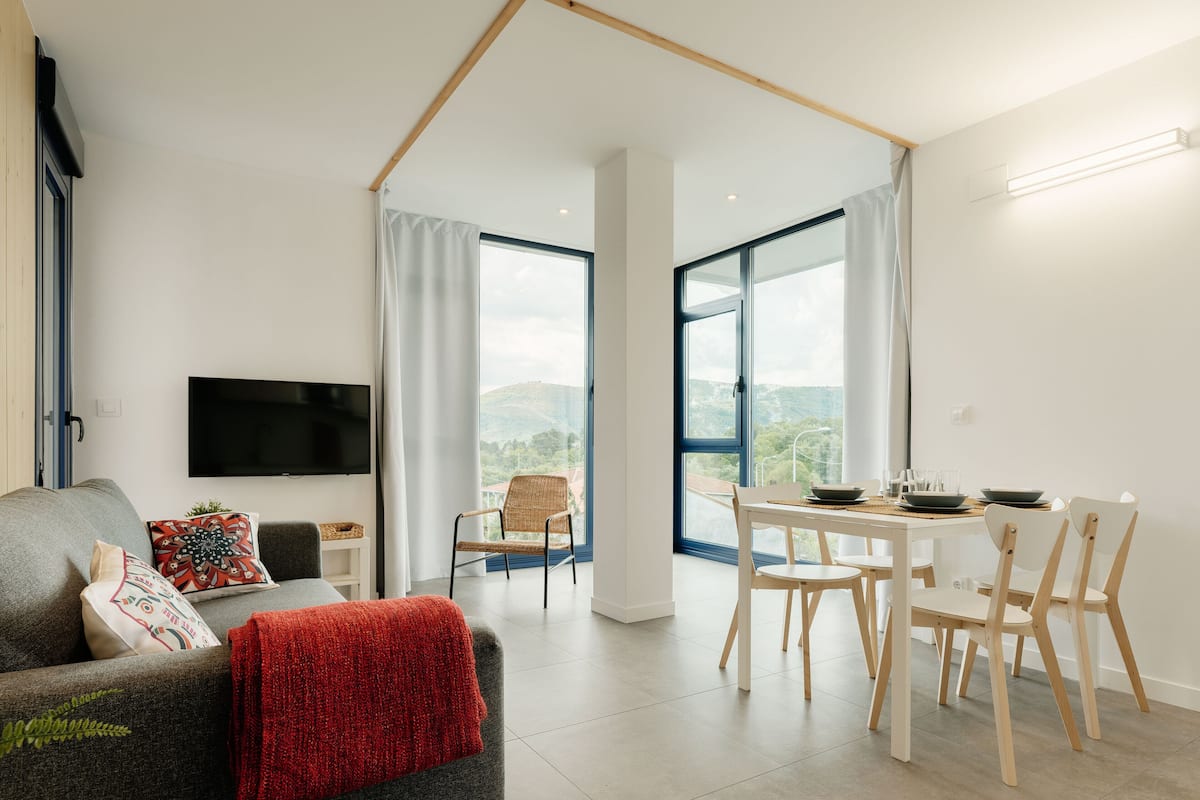 A modern living room with a flat-screen TV, a sofa, a dining table with chairs, and large windows offering a view of the outdoors.