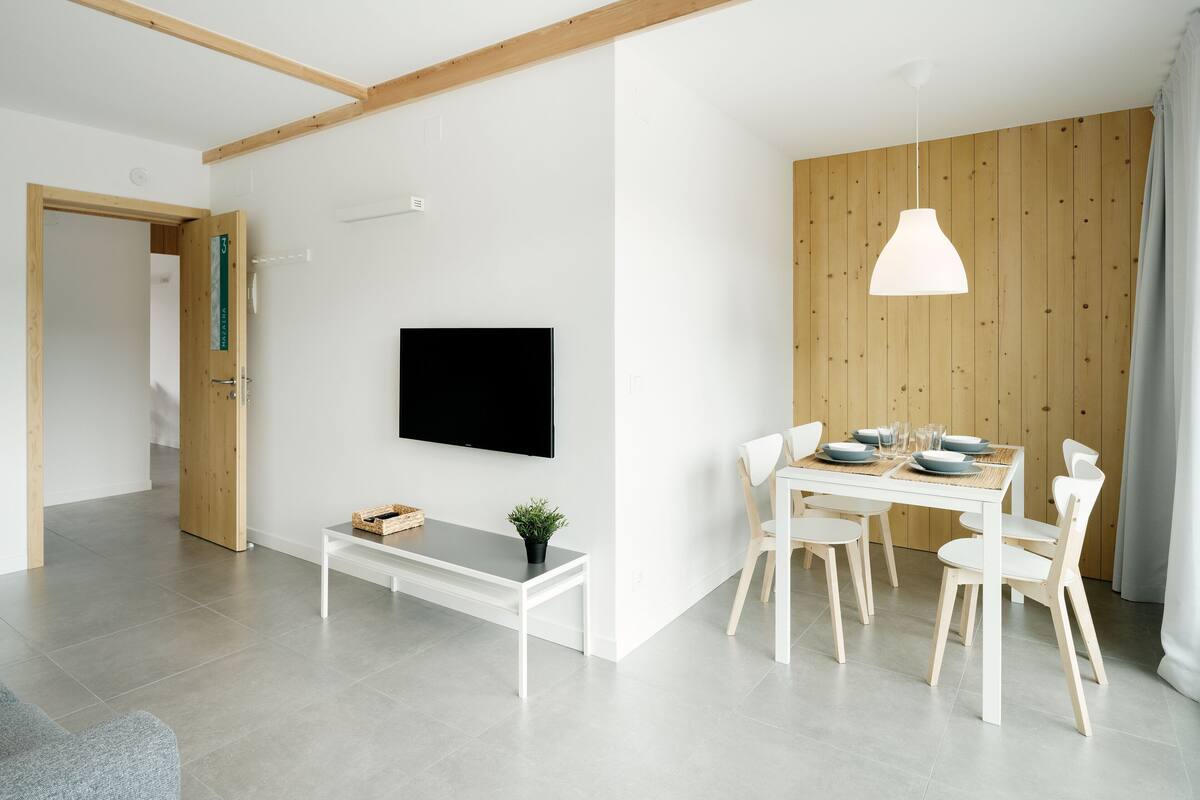 A modern living room with a flat-screen TV mounted on the wall, a dining table with chairs, and a bench.