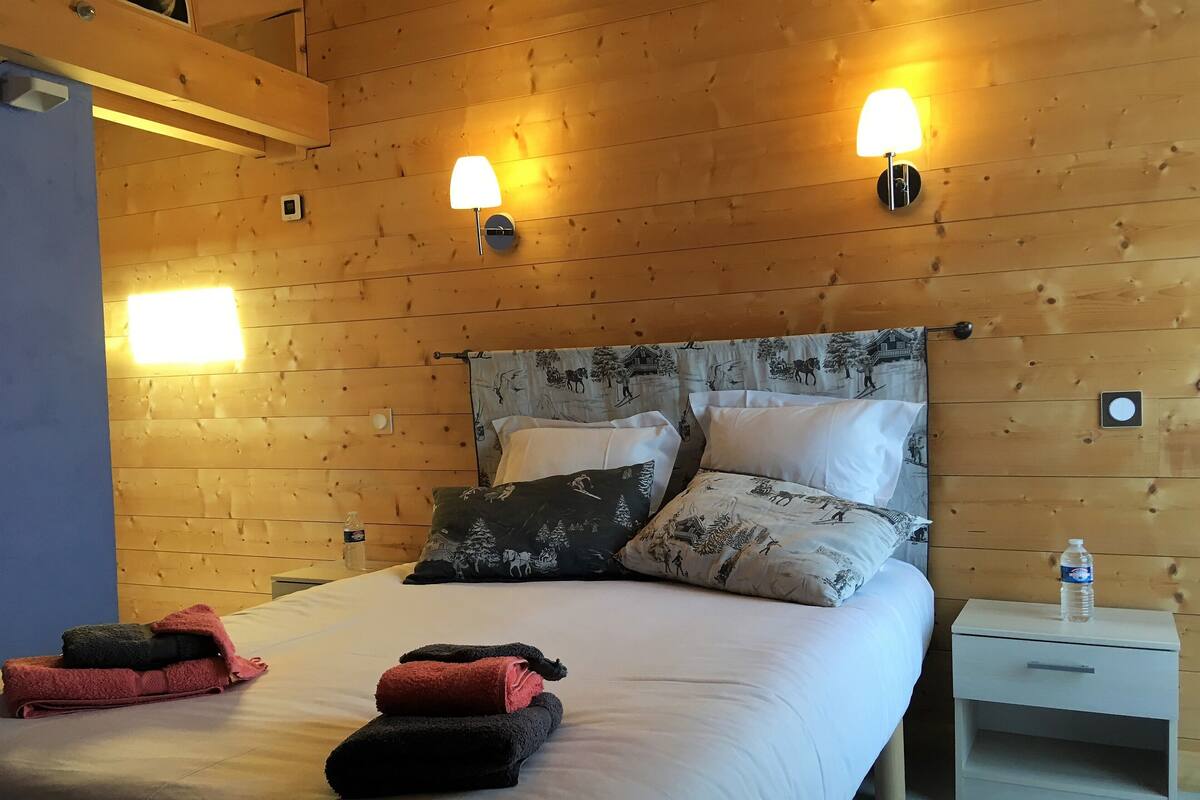 Une pièce aux murs lambrés, comprenant un lit, deux lampes fixées au mur et une table de chevet sur laquelle se trouve une bouteille.