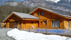 Eine Holzhütte mit geneigtem Dach, einer eingezäunten Terrassenfläche und Blick auf schneebedeckte Berge im Hintergrund.