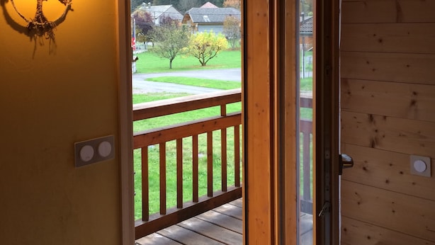 Eine Holztür führt zu einer Terrasse mit Geländer, einer wandmontierten Leuchte und Blick auf eine grüne Landschaft mit Bäumen und Häusern.