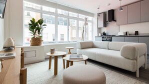 Ein modernes Wohnzimmer mit einer Couch, einem Couchtisch und einem Esstisch. Ein großes Fenster bietet einen Blick auf Gebäude außerhalb.