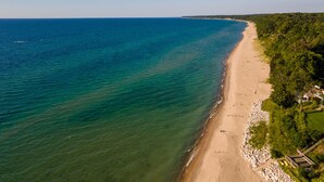 Une vue aérienne d’une plage aux eaux cristallines, bordée d’un rivage sablonneux et d’une zone boisée où se trouvent des bâtiments.