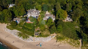 Vue aérienne d’une propriété côtière comprenant plusieurs maisons, une plage avec des parasols et une côte rocheuse.