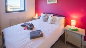 Ein ordentlich bezogenes Bett mit Kissen und Decke, ein Nachttisch mit Lampe und ein Fenster mit Blick auf Gebäude.