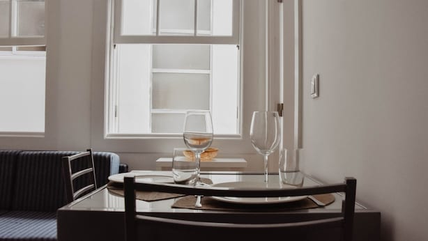 A dining area with a table set for two, a window with blinds, and a sofa in the background.