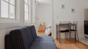A modern, minimalist room with a blue sofa, a wooden dining table with chairs, a small bedside table with a lamp, and a white door leading to another room.