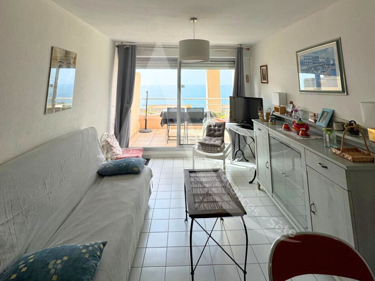 A living room with a sofa, a table, a TV, and a balcony with a view of the sea.