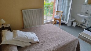 A bedroom with a bed, a chair, a radiator, a window with a view of greenery, and a bathroom with a sink and toilet. - Le Lithana (Pontorson)