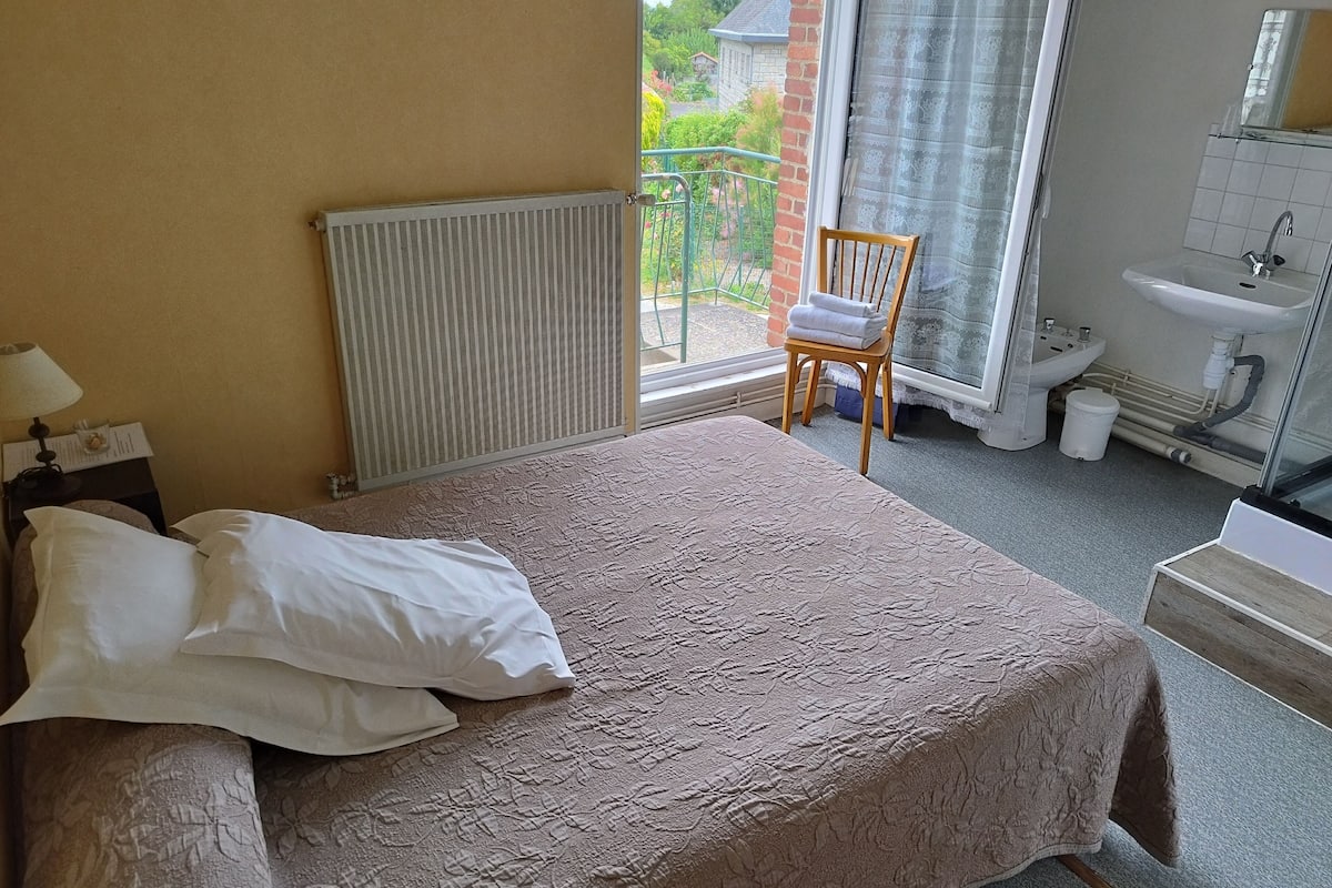 A bedroom with a bed, a chair, a radiator, a window with a view of greenery, and a bathroom with a sink and toilet.