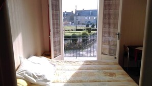 A bedroom with a bed, a window with curtains, and a view of a building and greenery outside. - Le Lithana (Pontorson)
