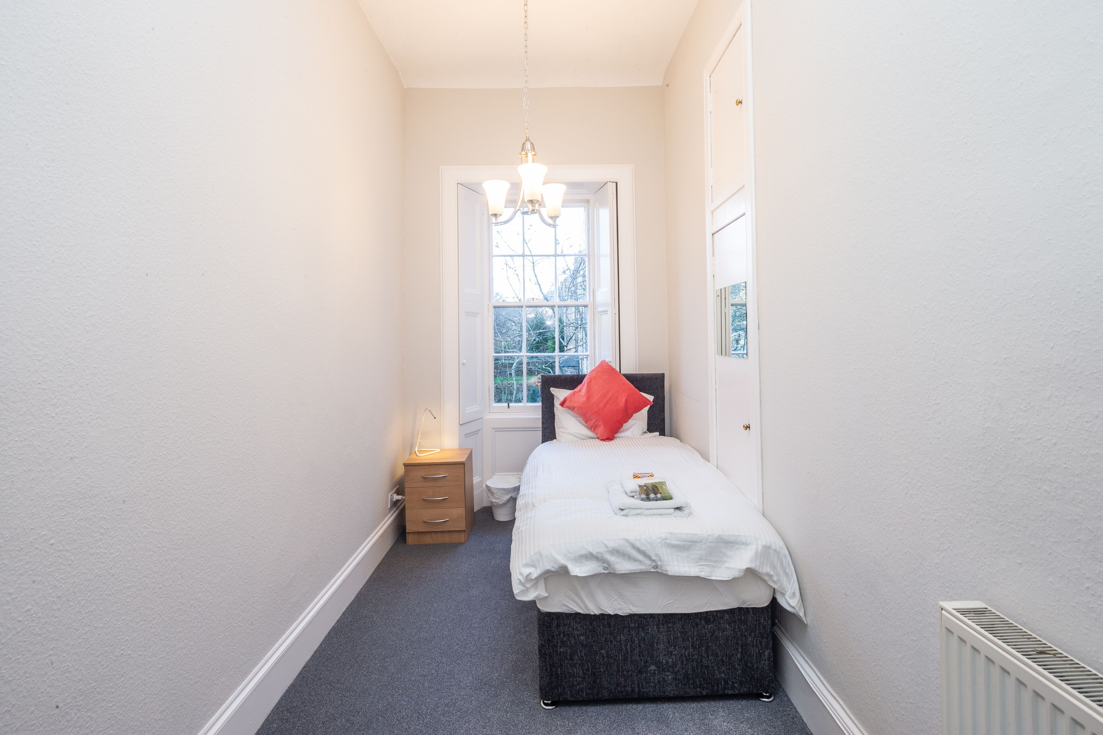 A narrow hallway leading to a bedroom with a bed, bedside table, and a window.