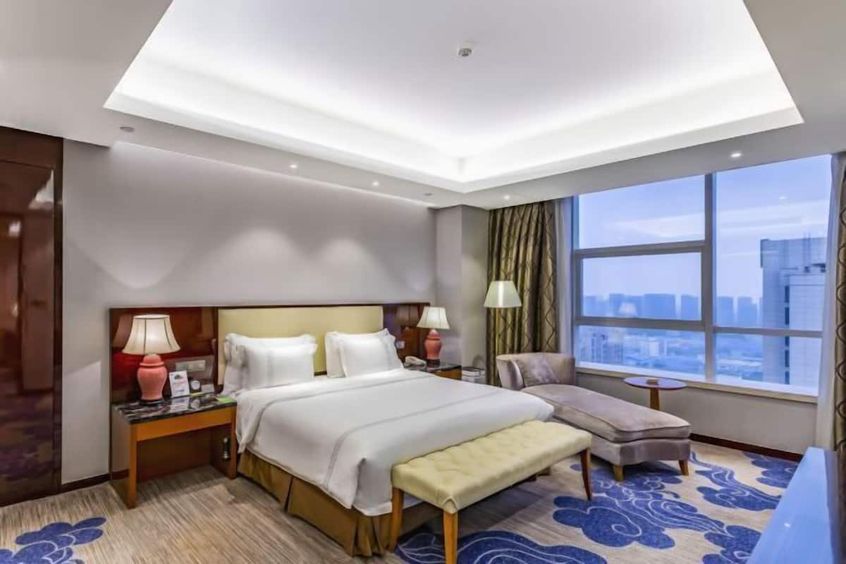 Een hotelkamer met een bed, nachtkastjes, een bankje, een raam met uitzicht op de stad en een plafondlamp.