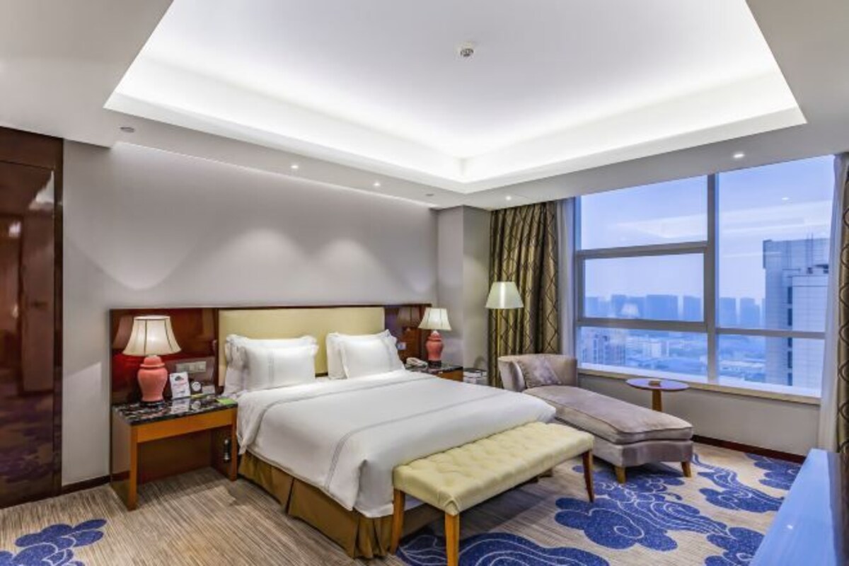Una habitación de hotel con cama, mesitas de noche, un banco, una ventana con vistas a la ciudad e iluminación en el techo.