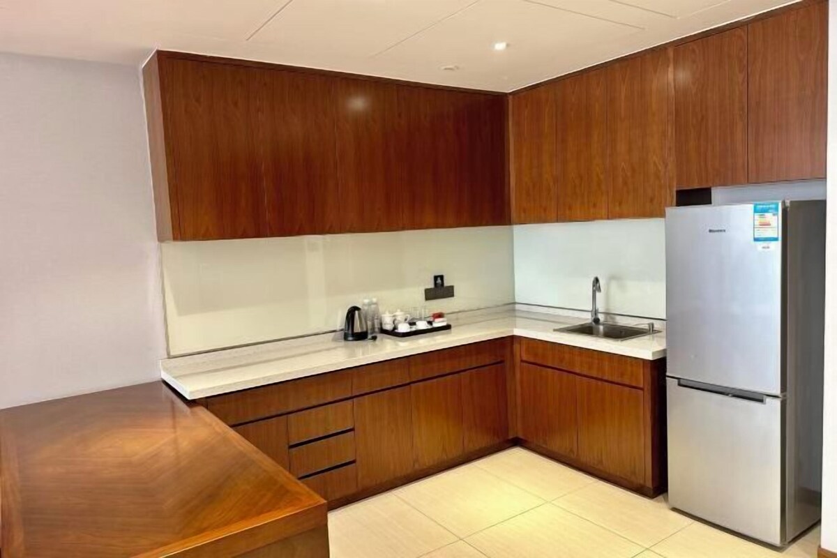 A kitchen with wooden cabinets, a stainless steel refrigerator, a sink, and a countertop with a kettle and cups.