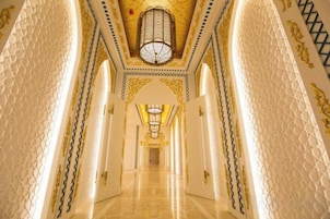 A corridor with ornate walls and a patterned ceiling, leading to a room with a chandelier.