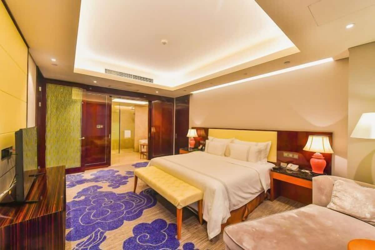 Een hotelkamer met een bed, nachtkastjes, een bankje, een televisie en een deur naar een andere kamer.