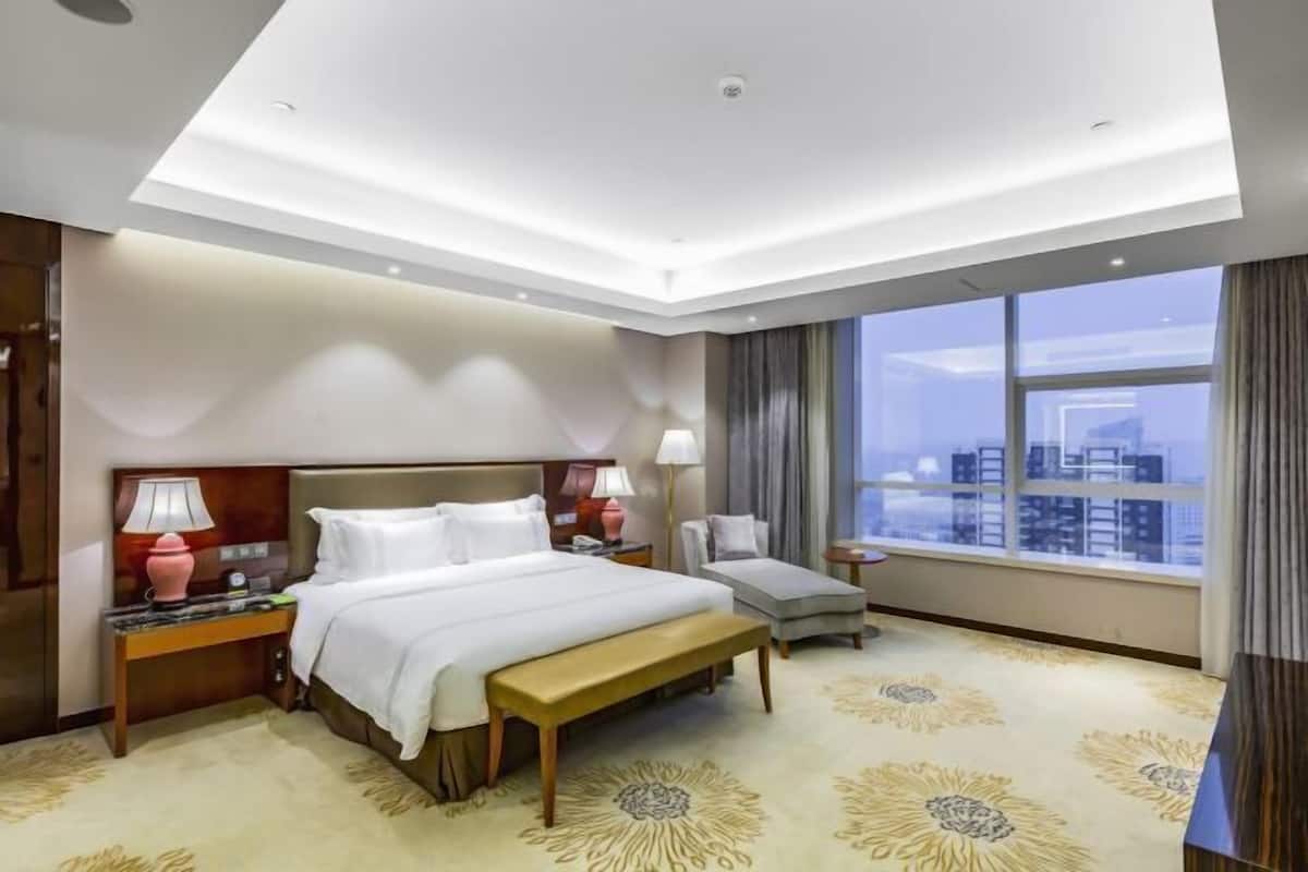 Een hotelkamer met een bed, nachtkastjes, een bankje, een stoel, een groot raam met uitzicht op de stad, en een kamerbreed tapijt met dessin.