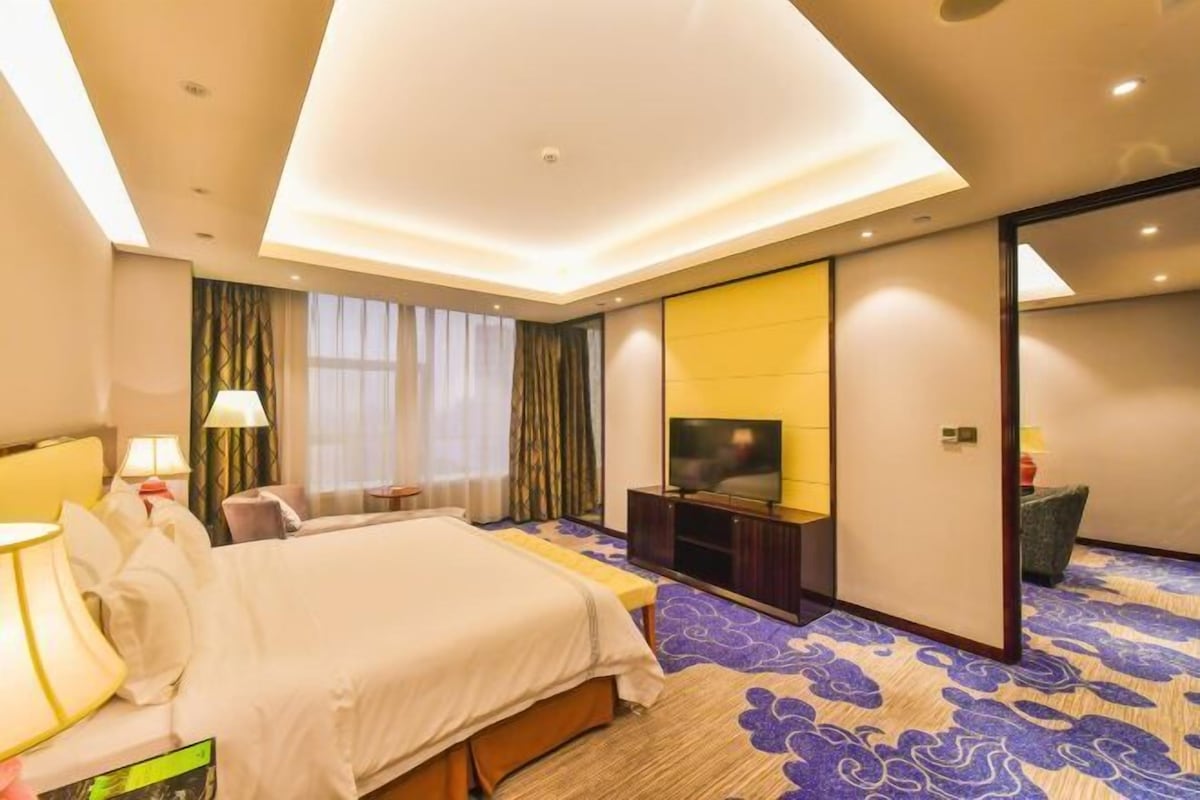 A hotel room with a bed, a TV, a desk, and a large window.