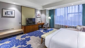 A hotel room with a bed, a desk with a TV, a chair, and a window with curtains.