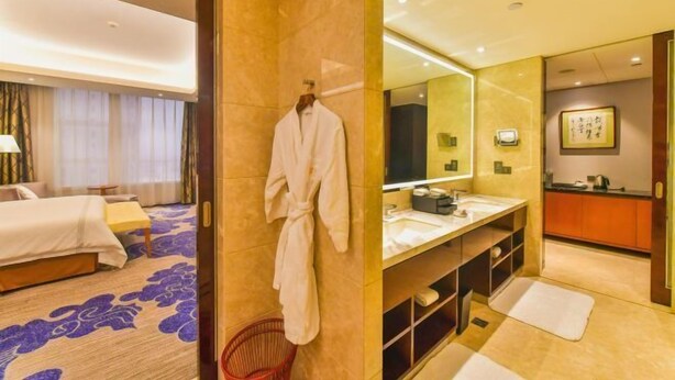 A hotel room with a bed, a robe hanging on the wall, a bathroom with a sink and mirror, and a small red basket.